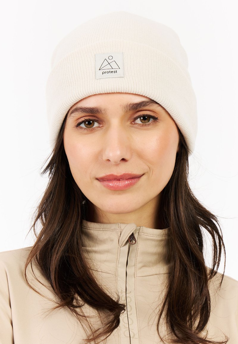 Woman with long brown hair wearing a beige zip-up jacket and a cream knit beanie with a "protest" logo patch.