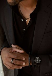 Black analog watch on wrist, dark gray suit jacket, black shirt with silver buttons, layered black bracelets, silver pendant necklace.