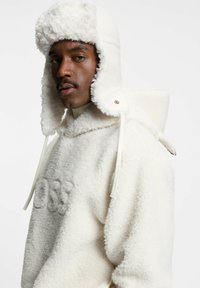 Felpa con cappuccio in pile bianco con finitura testurizzata e soffice. Presenta un cappuccio accogliente e paraorecchie, fissati con coulisse e dettagli in metallo dorato.