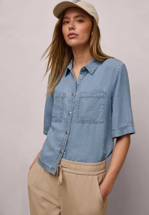 Frau mit langem blondem Haar, die eine beige Kappe, ein hellblaues Kurzarm-Hemd mit Knöpfen und beige Hose mit Kordelzug trägt, Hände in den Taschen.