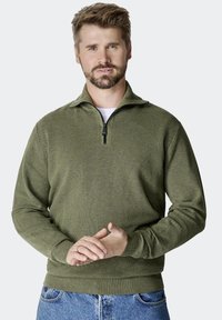 Oliwkowa bluza z zamkiem na szyi, z fakturowanej tkaniny, z prążkowanymi mankietami i dołem. Model nosi ją na białej koszuli, w komplecie z niebieskimi dżinsami.