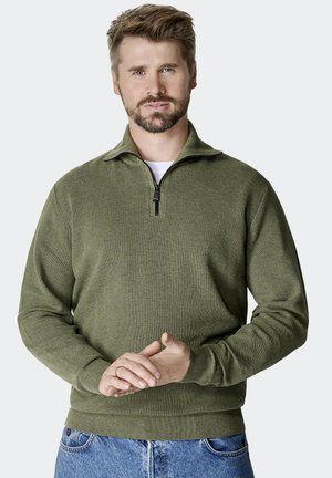 Olivgrüner Zip-Hoodie mit strukturiertem Stoff, gerippten Bündchen und Saum. Das Modell trägt ihn über einem weißen Hemd, kombiniert mit blauen Jeans.