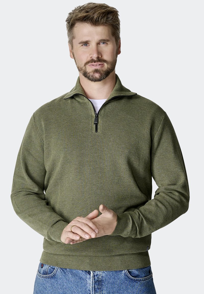 Oliwkowa bluza z zamkiem na szyi, z fakturowanej tkaniny, z prążkowanymi mankietami i dołem. Model nosi ją na białej koszuli, w komplecie z niebieskimi dżinsami.