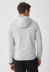 Sudadera de color gris claro con una textura suave, que cuenta con una capucha con cordón y un dobladillo acanalado. El modelo se muestra de espaldas.