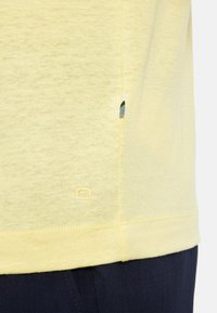 Geel t-shirt met korte mouwen van een lichte, gestructureerde stof, met een klein label met blauwe en zwarte accenten nabij de zijnaad.