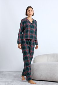 Zeleno-červený károvaný pyžamový set s gombíkovou blúzkou a nohavicami s elastickým pásom, vyrobený z mäkkej látky. Modelka stojí na svetlošedej koberci.