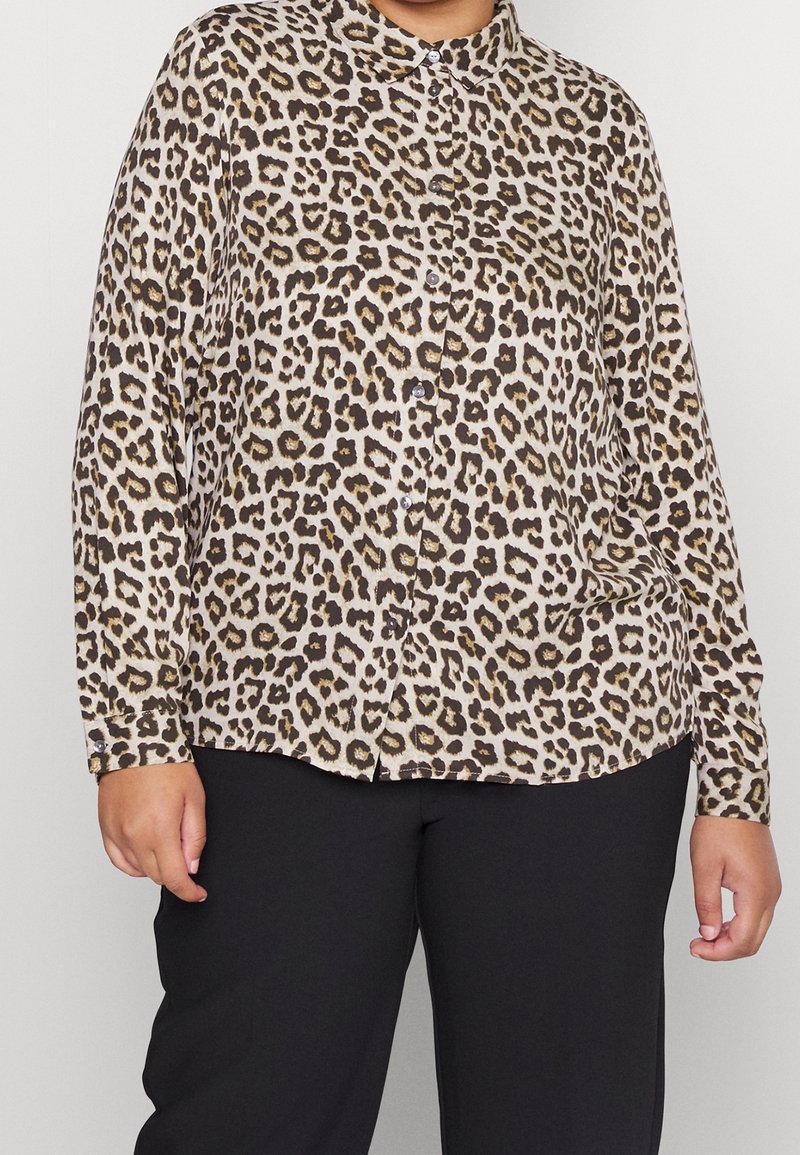 Chemise à manches longues boutonnée à imprimé léopard portée avec un pantalon noir sur une personne avec les mains relâchées le long du corps.