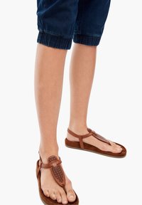 Bruna lädersandaler med en perforerad tårem och en platt, texturerad sula. Bärs med uppvikta marinblå byxor, vilket visar foten och benet.