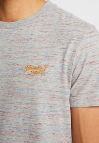 Detailní záběr osoby, která má na sobě fialově a šedě skvrnitý tričko s žlutým vyšitým logem "SuperDry" na hrudi.