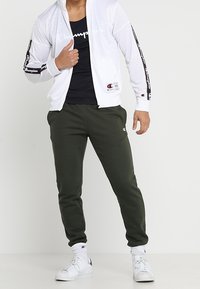 Vitt zip-up jacka med svart logotyp på ärmarna, svart linne, olivgröna sweatpants och vita sneakers med svarta detaljer.