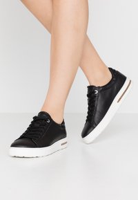 Baskets en cuir noir avec semelles en caoutchouc blanc, dotées de lacets ronds, d'œillets en argent et d'un accent texturé à l'arrière.