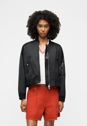 Mujer con cabello rizado que lleva una chaqueta bomber negra, camiseta blanca con cuello en v y rayas rojas, y pantalones cortos rojos de cintura alta con bajos deshilachados.