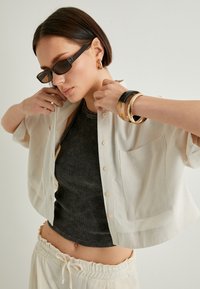 Camicetta a maniche corte color crema con bottoni sopra un top aderente scuro, abbinata a braccialetti a strati e occhiali da sole eleganti.