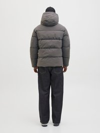 Person som bär en grå huvanpuffjacka, mörka vida jeans och en svart keps, stående vänd bort från kameran mot en enfärgad vit bakgrund.