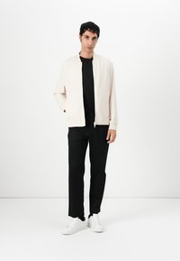 Light beige bomber jacket with a zip front, black t-shirt underneath, black trousers, and white sneakers, styled in a minimalist fashion.