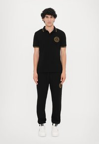 Cămașă polo neagră cu accente galbene și logo, asociată cu pantaloni jogger negri; prezintă o croială lejeră și un aspect casual.