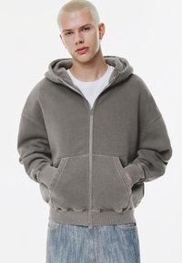 Hooded zip-up sweatshirt in gray. Features large front pockets, ribbed cuffs and hem, and soft, textured fabric. Worn over a white shirt.