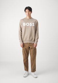 Sudadera beige con letras blancas en negrita "BOSS", combinada con pantalones cargo marrones con bolsillos laterales y zapatillas blancas. Diseño simple y casual.