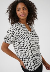 Blouse imprimée en noir et blanc avec un col en V, des manches courtes et un tissu texturé présentant un motif abstrait de formes.