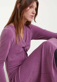 Vestido largo de punto morado con un detalle retorcido en la cintura, mangas largas y una textura suave. La tela tiene un ligero brillo.