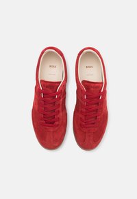 Red suede sneakers with a low-top design, featuring round laces, classic stitching, and a textured rubber sole. "BOSS" embossed on the side.