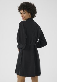 Vestido de mezclilla negra con mangas largas abullonadas, cuello fruncido y cintura ceñida. Presenta un dobladillo a la altura de la rodilla y bolsillos laterales.
