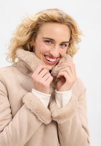 Beige Mantel mit Kunstfellkragen, weicher Textur, mit einem Verschluss und gepolsterten Bündchen. Das Modell hält den Kragen und zeigt dabei die Designdetails.