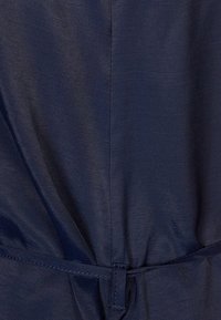 Mörkblått tyg, slät textur med en lätt lyster. Har en knytmidja och en subtil, samlad design. Inga synliga mönster.