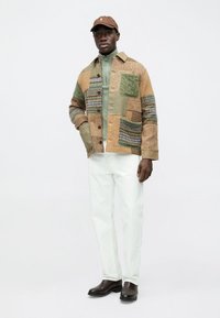 Casaco de retalhos castanho e verde com várias texturas e padrões, combinado com gola alta verde-claro, calças brancas e sapatos escuros.