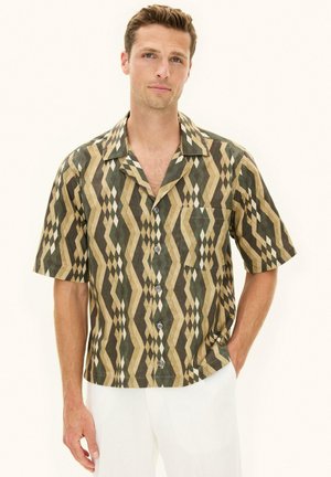 Mann trägt kurzärmliges Hemd mit braunem, beigem und grünem geometrischem Muster, kombiniert mit weißen Hosen, steht mit einer Hand in der Tasche.