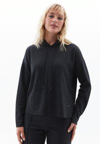 Tmavě šedá mikina z měkké tkaniny, která má kapuci, stahovací šňůrky a uvolněný střih. Jednoduchý design bez viditelných vzorů nebo log.