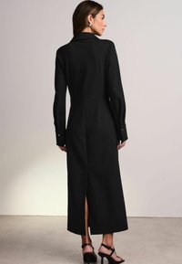 Sort langærmet kjole med tætsiddende liv, krave og slids bagpå. Glat stoftekstur med knaplukning ved håndleddene og minimalistisk design.