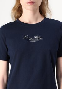 Tamno plavi pamučni majica s kratkim rukavima. Sadrži logo "Tommy Hilfiger New York City" u bijelom rukopisnom fontu preko prsa.