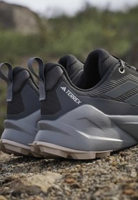 Paire de chaussures de trail Terrex grises et noires sur un sol rocheux avec languettes et semelles texturées visibles.
