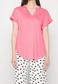 Kurzärmlige, pinke Bluse mit Henley-Ausschnitt und abgerundetem Saum. Der Stoff wirkt weich mit einer subtilen Textur. Kombiniert mit gepunkteten Hosen.