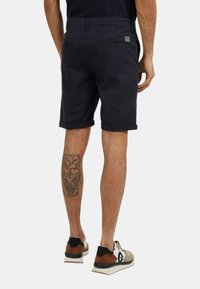 Pantaloni corti blu navy in cotone con orli arrotolati, due tasche posteriori e un piccolo etichetta del marchio, indossati con scarpe sportive multicolori.