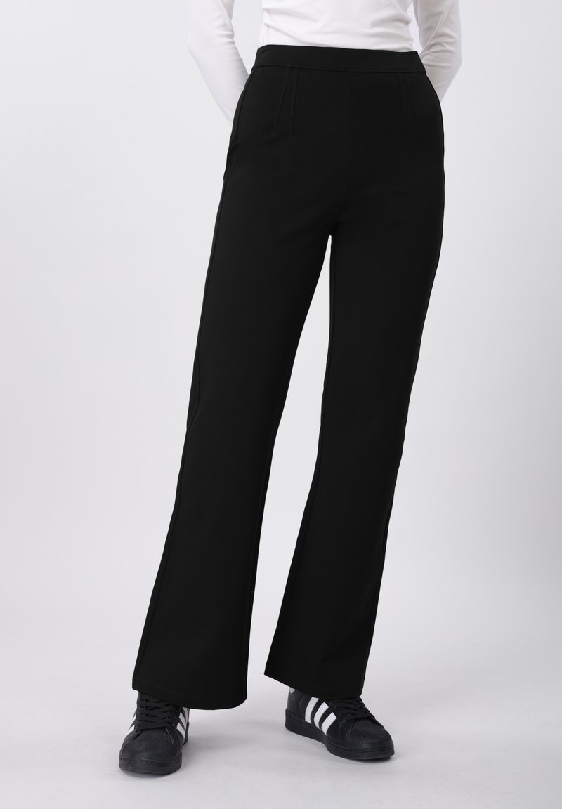 Pantalon noir évasé en matière lisse et extensible avec deux poches avant et une taille haute, porté avec des baskets noires.