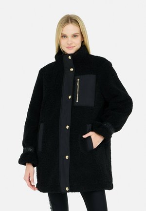 Veste en polaire noire avec un col haut, des accessoires dorés, une poche zippée et des panneaux en tissu contrasté. Coupe texturée et oversize avec des poches latérales.