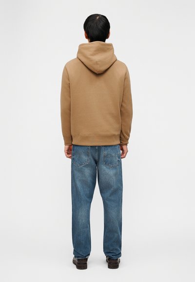 Άνδρας με την πλάτη γυρισμένη, φοράει μπεζ φούτερ με κουκούλα, μπλε τζιν άνετης γραμμής και σκούρα καφέ παπούτσια, μπροστά σε απλό λευκό φόντο.