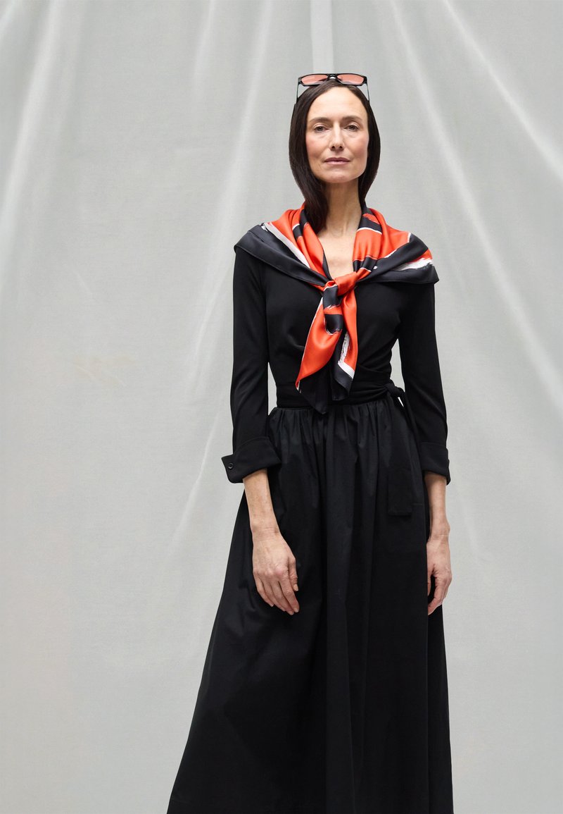 Juoda suknelė ilgomis rankovėmis, su juosmenį pabrėžiančia dalimi ir laisvai krintančia suknele, derinama su oranžinės ir juodos spalvų šilko skarele, rišama prie kaklo.