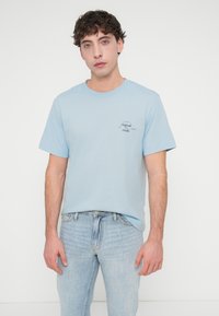 Ανοιχτό μπλε βαμβακερό t-shirt με κοντά μανίκια και στρογγυλή λαιμόκοψη, με ένα μικρό μαύρο γραφικό σχέδιο στο στήθος. Φορεμένο με ανοιχτόχρωμο τζιν.