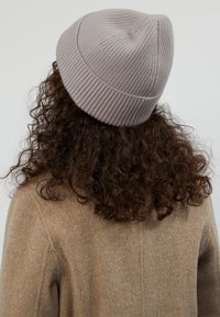 Vaaleanharmaa neulottu pipo, jossa on ribbikuvio ja tiukka istuvuus, käytetty kiharaisten hiusten päällä, yhdistettynä beigeen villakangastakkiin.