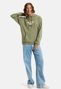Olivgrön hoodies med framficka, ribbade muddar och dragsko i huvan. Har en vit grafisk design på bröstet. Kombineras med ljusblå jeans.