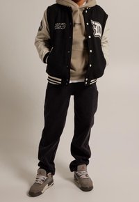 Giacca varsity nera e crema con dettagli ricamati, indossata sopra un felpa grigia. Pantaloni neri e sneakers grigie con accenti rossi.