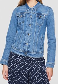 Chaqueta de mezclilla azul con cierre de botones, bolsillos en el pecho y mangas largas, llevada sobre pantalones de patrón azul marino.