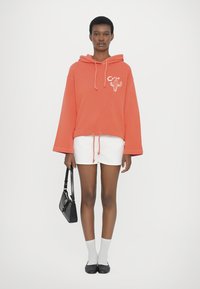 Korallefarbener oversized Hoodie mit Druck eines Cactus auf der Vorderseite, Kordelzugsaum, kombiniert mit weißen Shorts. Die Accessoires umfassen eine kleine schwarze Handtasche und Knöchelsocken.