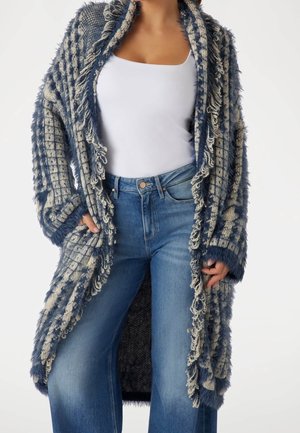 Femme portant un cardigan en maille bleu et beige à franges, par-dessus un débardeur blanc et un jean bleu taille haute à jambes larges.