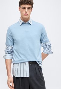 Νεαρός άνδρας που φοράει ανοιχτό μπλε κοντομάνικο πουκάμισο πάνω από ριγέ μακρυμάνικο πουκάμισο, συνδυασμένο με σκούρο παντελόνι και μαύρη ζώνη.