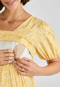 Frau in gelbem Blumenkleid hebt die Oberseite an, um ein beiges Brustpumpen-Bh zu zeigen, während ihre Hände einen Ring und eine Halskette präsentieren.