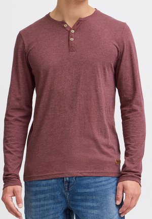 Uomo che indossa una maglietta Henley bordeaux a maniche lunghe con tre bottoni e jeans blu, in piedi davanti a uno sfondo semplice.